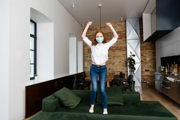 Frau in medizinischer Maske zeigt Yah-Geste beim Sprung auf Couch — Stockfoto