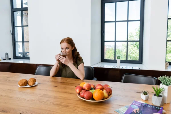 Frau trinkt Kaffee in der Nähe von Croissants, Obst und Zeitschriften auf dem Tisch — Stockfoto