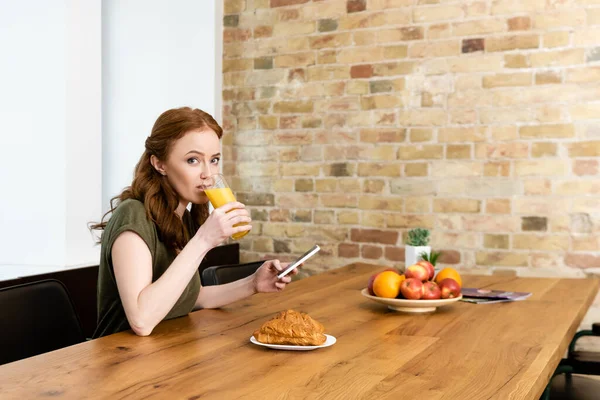 Mujer mirando a la cámara mientras usa el teléfono inteligente y bebe jugo de naranja en casa - foto de stock