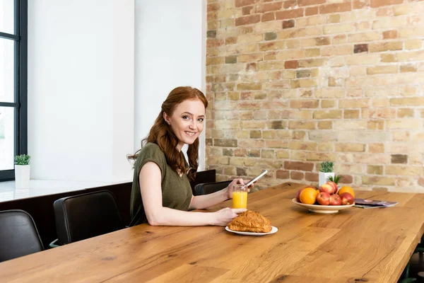 Улыбающаяся женщина смотрит в камеру во время использования смартфона возле завтрака на столе — стоковое фото