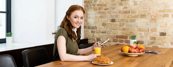 Horizontal crop of smiling woman using smartphone near croissants and orange juice on table — Stock Photo
