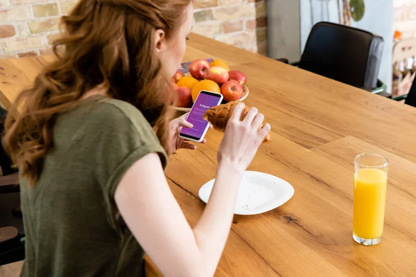 KYIV, UCRAINA - 8 MAGGIO 2020: Vista laterale della donna che utilizza lo smartphone con app instagram e tiene il croissant a tavola — Foto stock