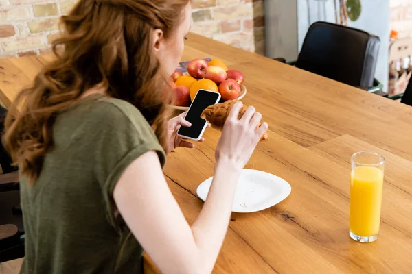 Selective focus of woman using smartphone while holding croissant near glass of orange juice on table — Stock Photo