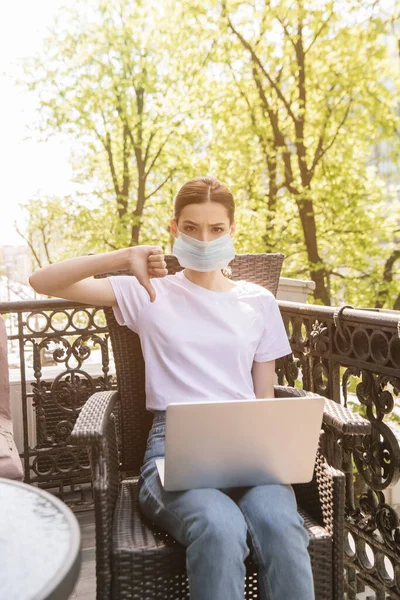 Joven freelancer en máscara médica sentado en silla con portátil y mostrando el pulgar hacia abajo - foto de stock