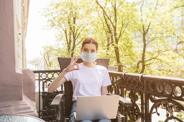 Giovane freelance in maschera medica seduto sulla sedia con computer portatile e mostrando segno ok — Foto stock