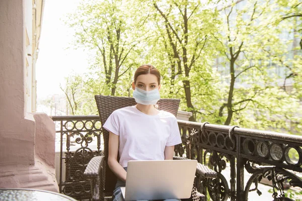 Фрилансер в медицинской маске сидит на стуле и пользуется ноутбуком на балконе — стоковое фото