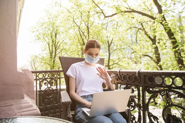 Mädchen in medizinischer Maske sitzt auf Stuhl und winkt mit der Hand beim Videoanruf auf dem Balkon — Stockfoto