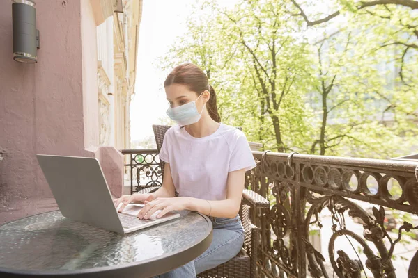 Junge Freiberuflerin in medizinischer Maske sitzt auf Stuhl und benutzt Laptop auf Balkon — Stockfoto