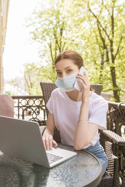 Jovem freelancer em máscara médica falando no smartphone e usando laptop na varanda — Fotografia de Stock