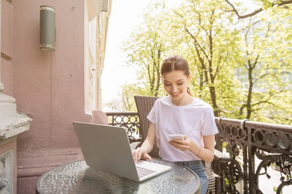 Feliz y atractivo freelancer mirando el teléfono inteligente cerca de la computadora portátil en la mesa - foto de stock