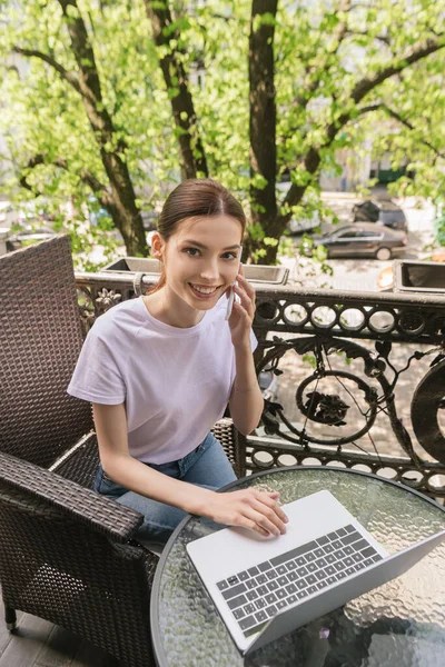 Freelancer alegre e atraente falando no smartphone perto do laptop na mesa — Fotografia de Stock
