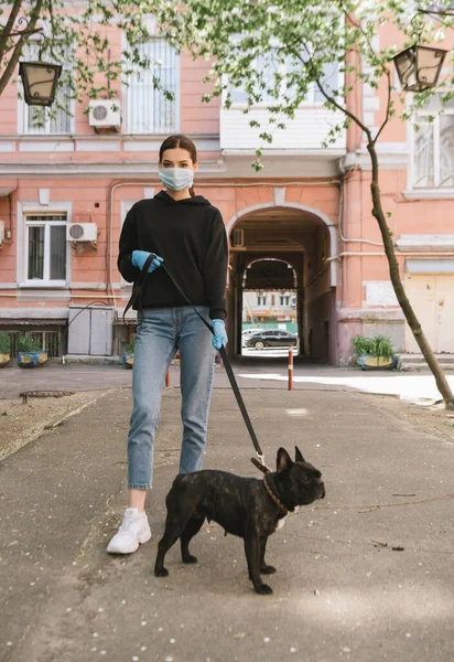 Jovem mulher em máscara médica e luvas de látex de pé com bulldog francês na rua — Fotografia de Stock