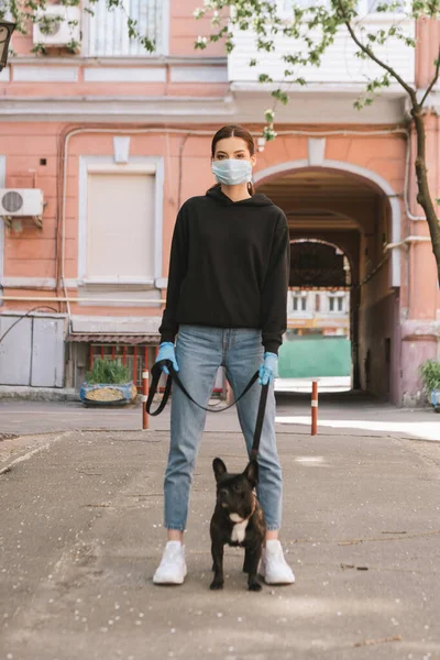 Mujer joven en máscara médica y guantes de látex de pie con bulldog francés lindo en la calle - foto de stock