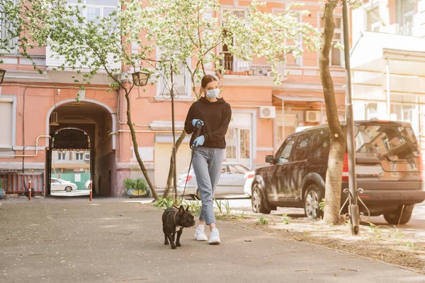 Mujer joven en máscara médica caminando con bulldog francés lindo en la calle - foto de stock