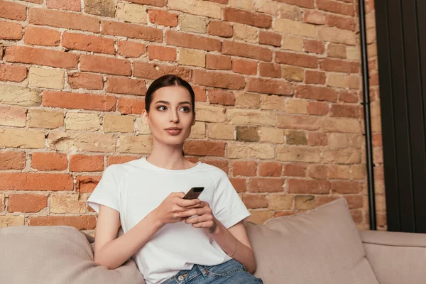 Attraente giovane donna seduta sul divano e tenendo telecomando — Foto stock