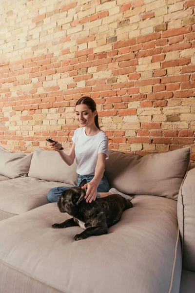 Feliz joven mujer sosteniendo mando a distancia y sentado en el sofá con bulldog francés - foto de stock