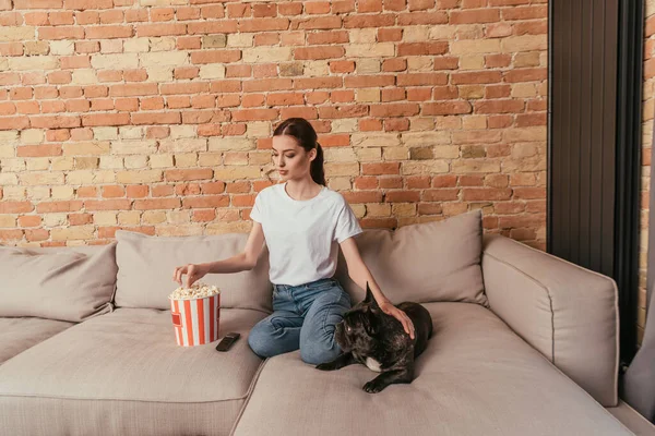 Щаслива жінка добирається до попкорна і сидить на дивані з милим французом бульдогом — стокове фото