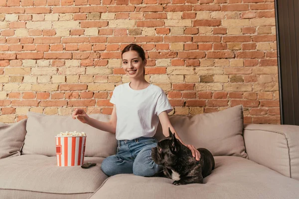 Щаслива жінка тримає попкорн і сидить на дивані з милим французьким бульдогом — стокове фото