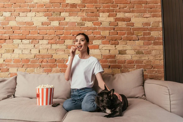 Щаслива жінка їсть попкорн і сидить на дивані з милим французьким бульдогом під час перегляду фільму — стокове фото