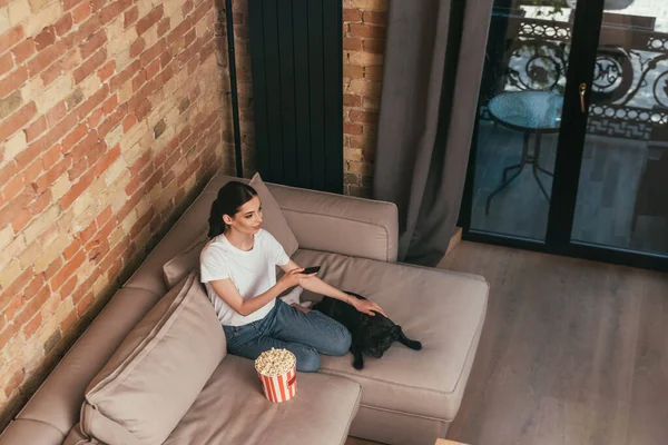 Вид зверху весела жінка сидить на дивані біля відра з попкорном і милий французький бульдог, тримаючи пульт дистанційного керування — стокове фото