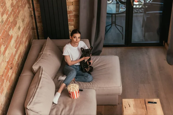 Vue aérienne d'une jolie fille assise sur un canapé près d'un seau de maïs soufflé et d'un joli bouledogue français — Photo de stock