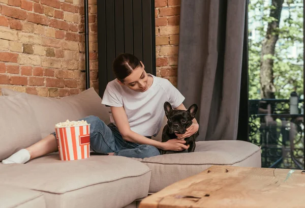 Messa a fuoco selettiva di attraente ragazza seduta sul divano vicino popcorn secchio e toccando carino bulldog francese — Foto stock