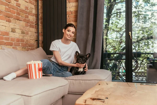 Enfoque selectivo de chica alegre sentado en el sofá cerca de palomitas de maíz cubo y tocando lindo bulldog francés - foto de stock