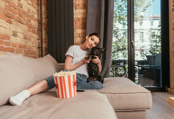 Веселая девушка сидит на диване возле ведра с попкорном и обнимает милого французского бульдога — стоковое фото