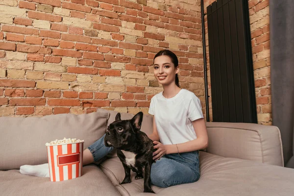 Lächelnde Frau sitzt auf Sofa neben Popcorn-Eimer und berührt niedliche französische Bulldogge — Stockfoto
