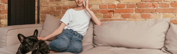 Panoramic shot of girl sitting on sofa and talking on smartphone while touching cute french bulldog — Stock Photo