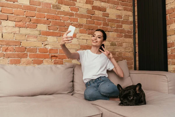 Glückliche Frau zeigt Friedenszeichen, während sie Selfie mit süßer französischer Bulldogge im Wohnzimmer macht — Stockfoto