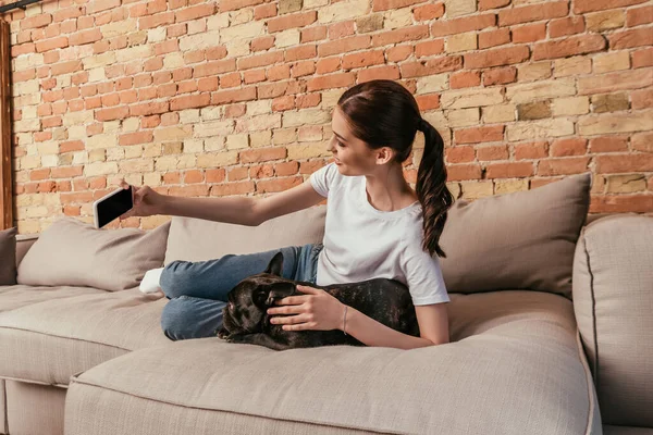Sonriente joven mujer tomando selfie con lindo bulldog francés en sala de estar - foto de stock