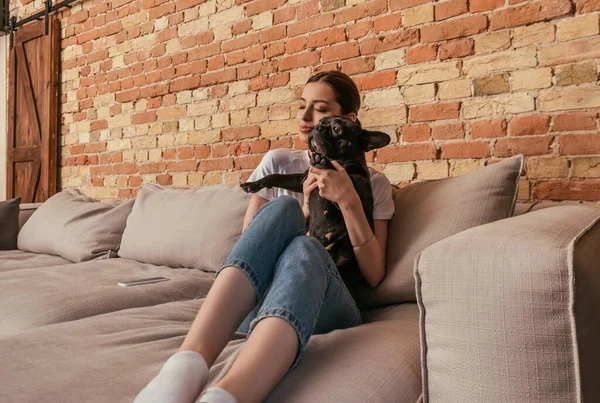Feliz chica sosteniendo en brazos lindo bulldog francés mientras está sentado en el sofá cerca de teléfono inteligente - foto de stock