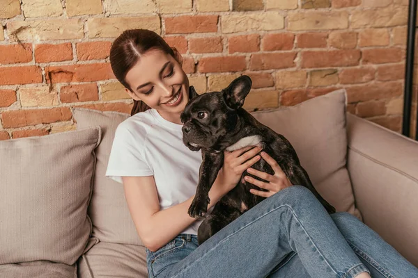 Happy girl holding in arms cute french bulldog while sitting on sofa — Stock Photo