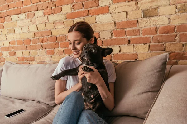 Chica feliz sosteniendo en brazos lindo bulldog francés mientras está sentado en el sofá cerca de teléfono inteligente con pantalla en blanco - foto de stock