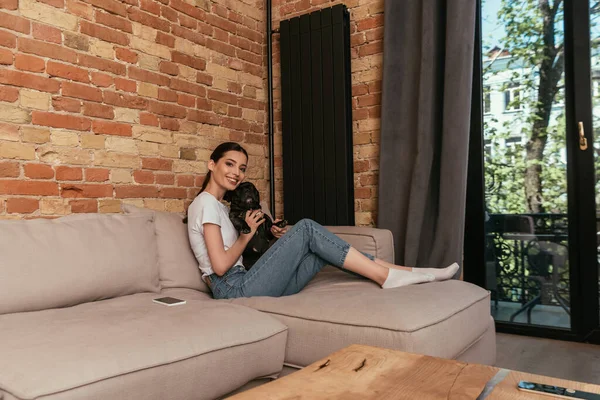 Cheerful girl holding in arms cute french bulldog while sitting on sofa near smartphone — Stock Photo