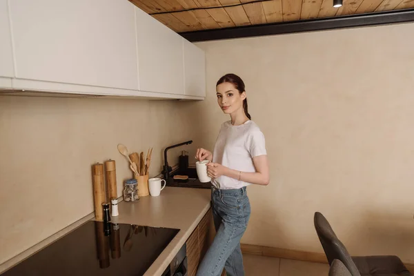 Atractiva mujer joven sosteniendo taza de café en la cocina - foto de stock