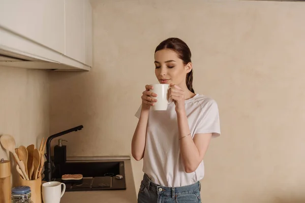 Atractiva chica oliendo café mientras sostiene la taza en la cocina - foto de stock