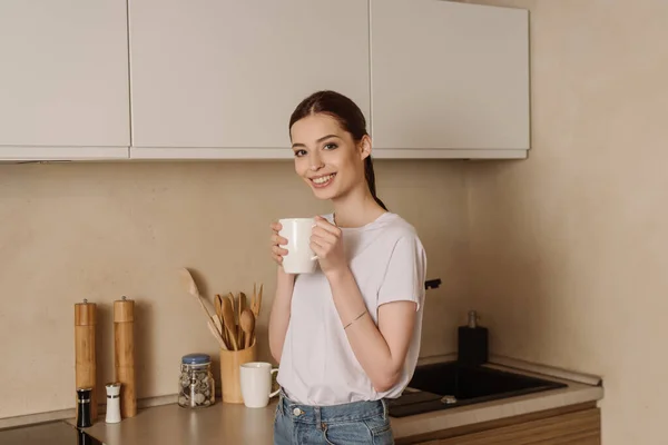 Feliz joven mujer sosteniendo la taza de café en la cocina - foto de stock