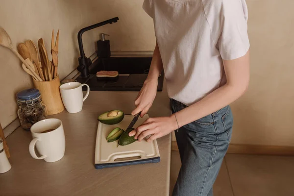 Vue recadrée de la femme tenant couteau tout en coupant l'avocat savoureux près des tasses — Photo de stock