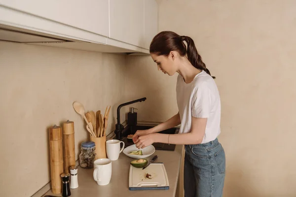 Jeune femme mettant l'avocat savoureux dans un bol près de la planche à découper et couteau — Photo de stock