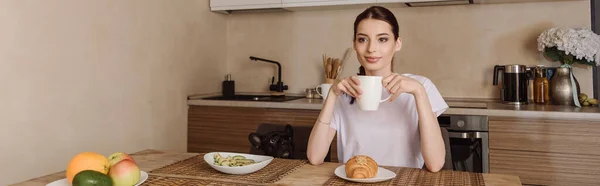 Horizontales Bild einer attraktiven Frau, die eine Tasse Kaffee in der Nähe von leckerem Frühstück und französischer Bulldogge hält — Stockfoto