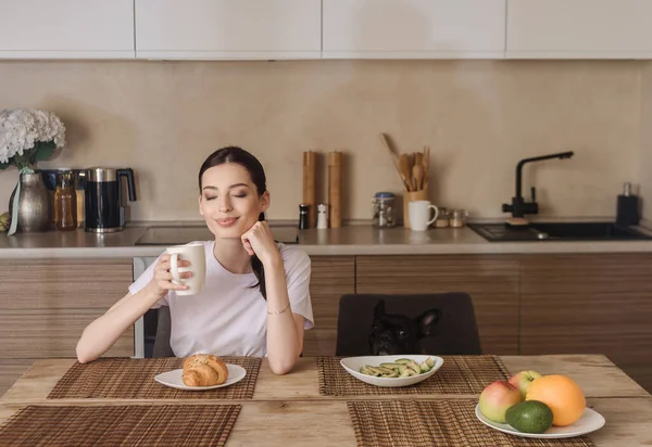 Femme heureuse tenant tasse de café près de savoureux petit déjeuner et bulldog français — Photo de stock