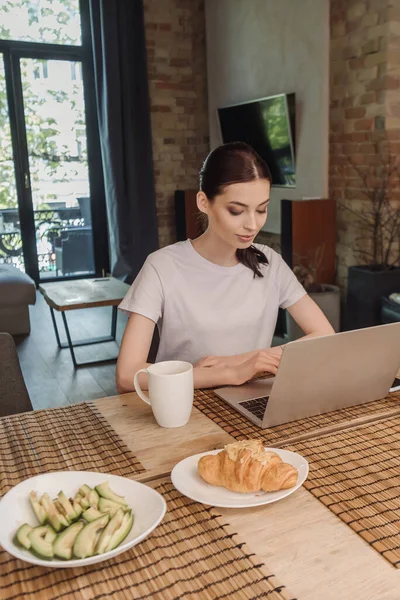 Atractivo freelancer utilizando el ordenador portátil cerca del desayuno en la mesa - foto de stock