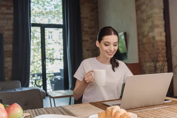 Щасливий фрілансер тримає чашку кави біля ноутбука і круасанів на столі — стокове фото