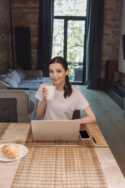 Happy freelancer holding cup of coffee near laptop, smartphone with blank screen and croissant on table — Stock Photo