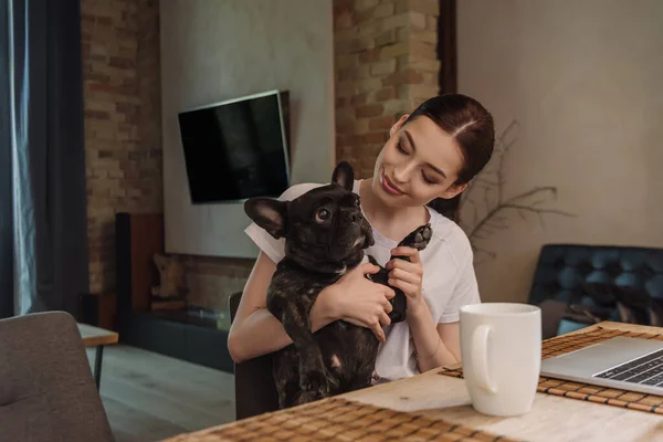 Alegre freelancer sosteniendo en brazos bulldog francés cerca de portátil y taza - foto de stock