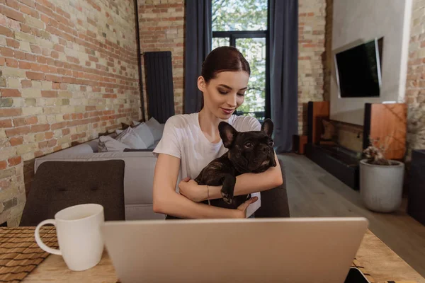 Selective focus of attractive freelancer holding in arms french bulldog near laptop and cup — Stock Photo