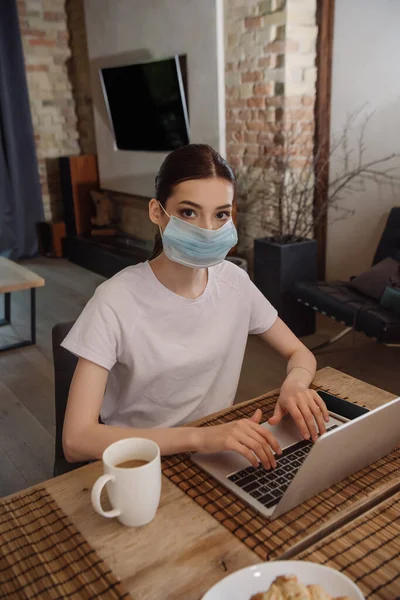 Joven freelancer en máscara médica escribiendo en el portátil cerca de la taza de café - foto de stock