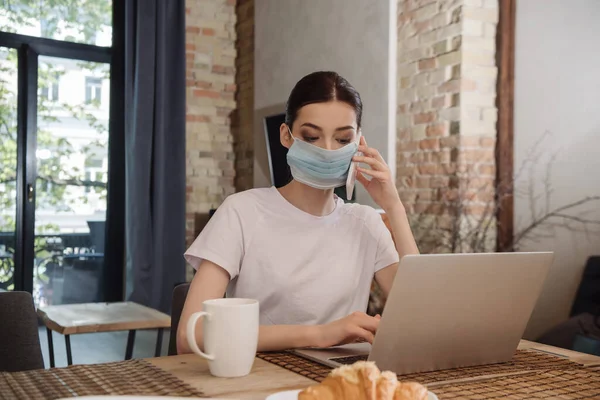 Junge Freiberuflerin in medizinischer Maske spricht auf Smartphone neben Laptop — Stockfoto
